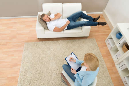 Patient Lying On Couch In Front Of Psychiatrist Writing On Clipboardの写真素材