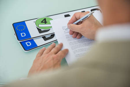 Close-up Of A Businessman Filling Car Sale Contract Form With Vehicle Registration Plate On Desk. Contract Paper Contains Placeholder Textの写真素材