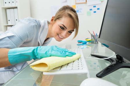 Portrait Of Young Happy Maid Cleaning Keyboard At Desk In Office