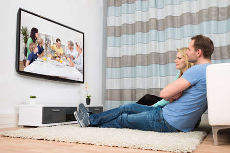 Couple Sitting On Carpet Watching Television In Living Room