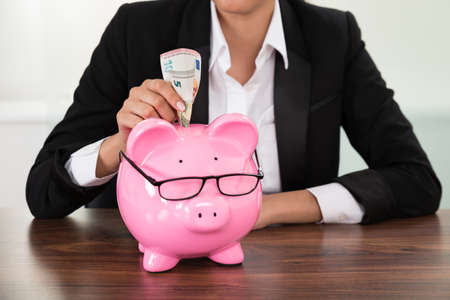Young Businesswoman Inserting Banknote In Piggybank At Deskの写真素材