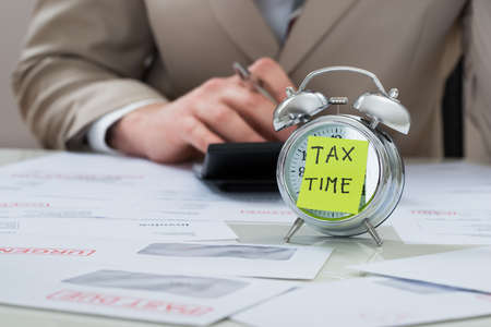 Close-up Of Businessman With Tax Time Reminder Note On Alarm Clock At Desk