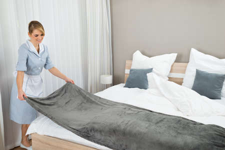 Happy Female Housekeeping Worker Making Bed In Hotel Roomの写真素材