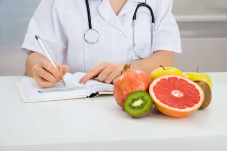 Close-up Of Female Dietician Writing Prescription With Fruits On Deskの写真素材