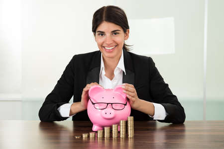 Young Happy Businesswoman With Piggybank And Stacks Of Coins At Deskの写真素材