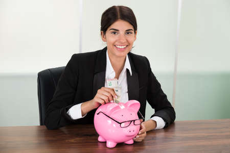 Young Businesswoman Inserting Banknote In Piggybank At Deskの写真素材