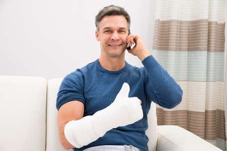 Man With Fractured Hand Sitting On Sofa Talking On Cellphoneの写真素材