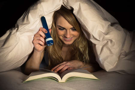 Young Woman Under Bedding Reading Book Holding Torchの写真素材