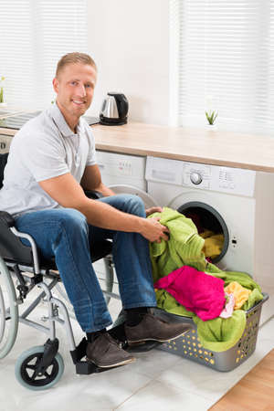 Young Happy Man On Wheelchair Putting Clothes Into The Washing Machineの写真素材