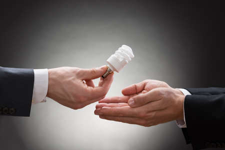 Close-up Of Businessperson Hand Offering Fluorescent Light Bulb To Other Businesspersonの写真素材
