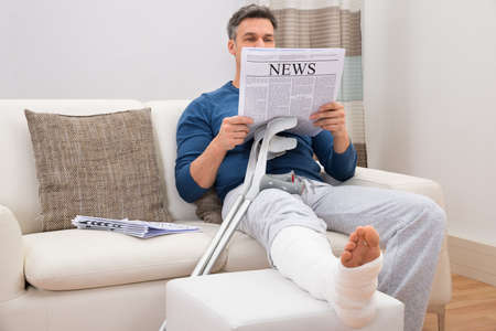 Disabled Man With Crutches Sitting On Sofa Reading Newspaperの写真素材
