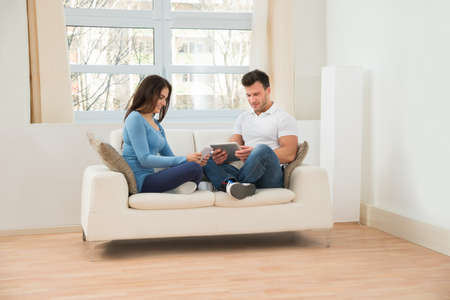 Smiling Young Couple Sitting On Couch Using Digital Tabletの写真素材