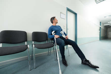 Mid-adult Man With Crutches Sitting On Chair In Hospitalの写真素材