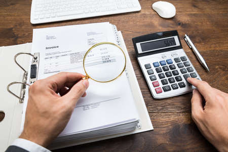 Close-up Of Businessperson Hand Examining Bills With Magnifying Glass At Desk