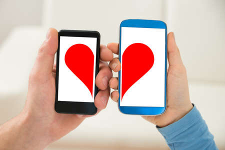Close-up Of A Couple's Hand Holding Cellphone With Half Red Heart Symbolの写真素材