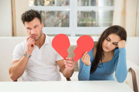 Portrait Of A Sad Couple Sitting On Chair Holding Red Broken Heart