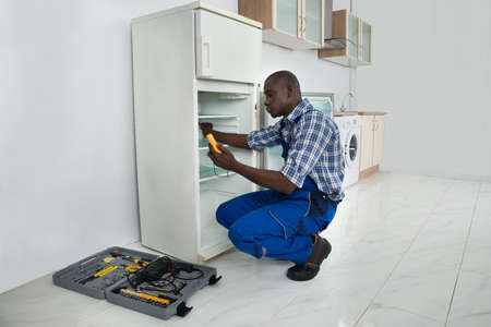 Young African Repairman Repairing Refrigerator In Kitchen Roomの写真素材