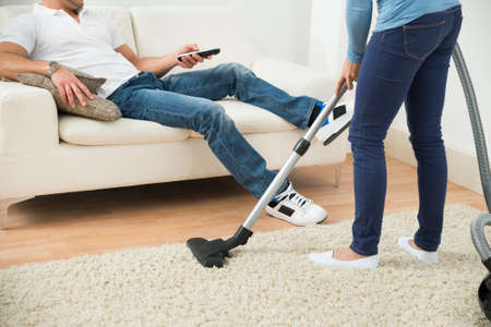 Close-up Of A Woman Cleaning Carpet In Front Of Man Sitting On Couchの写真素材
