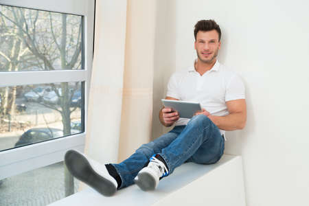Portrait Of A Smiling Young Man Sitting Near Window Holding Digital Tabletの写真素材