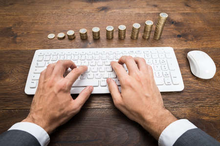 Close-up Of Businessman's Hand Using Keyboard In Front Of Stacked Coinsの写真素材