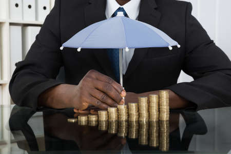 Close-up Of Businessman Protecting Stack Of Coins With Umbrella At Desk