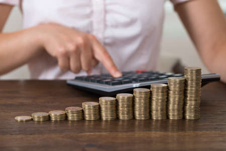 Close-up Of Businesswoman Calculating With Calculator In Front Of Stacked Coins At Deskの写真素材
