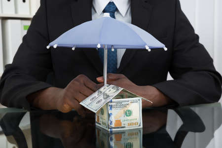 Close-up Of Businessman Hands Protecting House Of Banknote With Umbrella At Desk