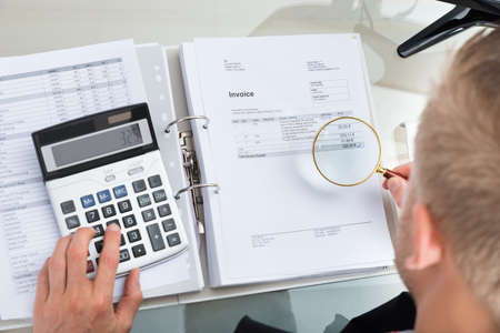 Close-up Of Businessman Inspecting Bills With Magnifying Glass While Using Calculator