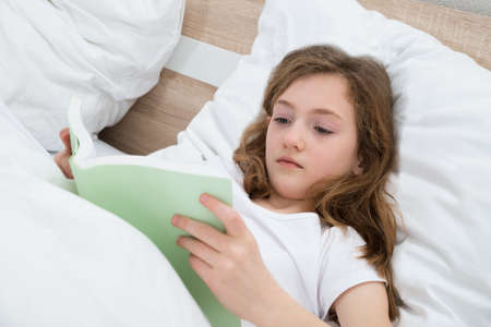 Cute Girl With Pillow Reading Book And Lying In Bed