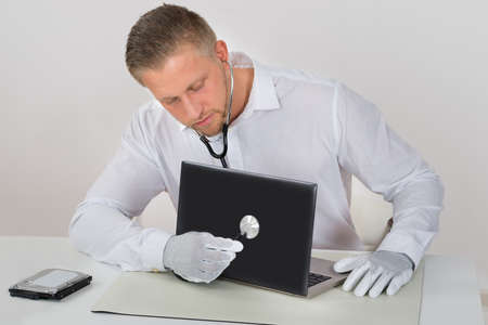 Young Male Technician Examining Laptop With Stethoscope At Deskの写真素材