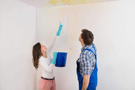 Young Woman With Bucket Showing Water Leaking From Damage Ceiling To Maintenance Guy
