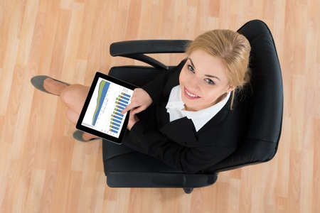 Happy Businesswoman Sitting On Office Chair With Digital Tablet Showing Graphsの写真素材