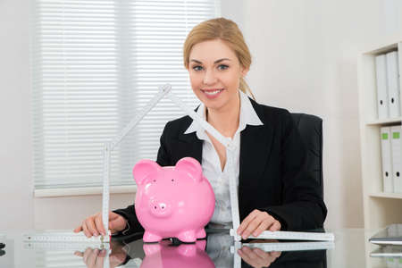 Happy Businesswoman Holding Ruler Folded In House Shape With Piggy Bank At Deskの写真素材