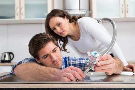 Young Woman Looking At Plumber Fixing Steel Tap In Kitchen Sinkの写真素材