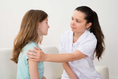 Young Female Doctor Comforting Girl Sitting On Sofa In Hospitalの写真素材