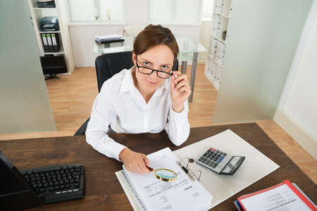 Young Businesswoman With Invoice And Magnifying Glass At Desk In Officeの写真素材