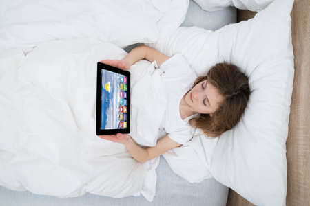 High Angle View Of Girl In Bed Holding Digital Tablet With Multi-colored Apps On Screenの写真素材