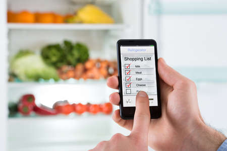 Close-up Of Person Hands Marking Shopping List On Mobile Phone In Front Open Fridgeの写真素材