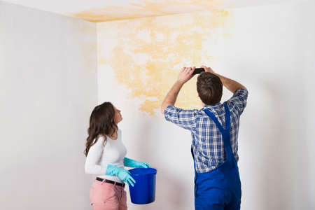 Handyman With Woman Photographing Water Damaged Ceiling With Mobile Phone At Home