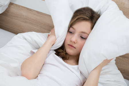 Girl On Bed Covering Ears With Pillow In Bedroomの写真素材