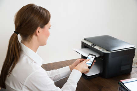 Young Businesswoman With Mobile Phone Showing Graph Connected To Printer At Deskの写真素材