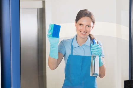 Young Happy Female Janitor Cleaning Glass With Detergent Spray Bottle And Sponge