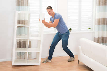 Young Man Moving Shelf In Living Roomの写真素材