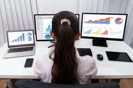 Rear View Of Businesswoman Analyzing Graphs On Multiple Computers At Deskの写真素材
