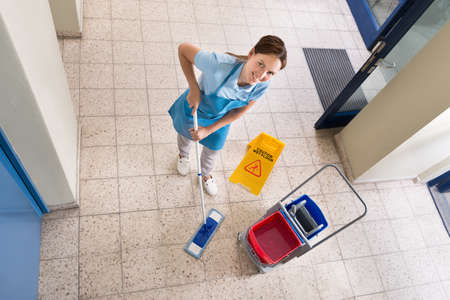 Happy Female Janitor Holding Mop With Cleaning Equipments And Wet Floor Sign In Corridorの写真素材