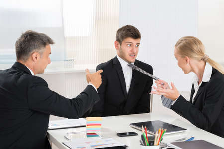 Two Businesspeople Quarreling To Young Businessman At Desk In Office