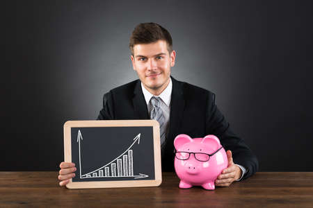 Young Businessman With Piggybank And Small Blackboard Showing Graph At Deskの写真素材