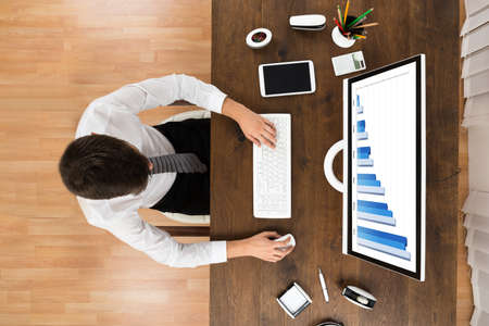 High Angle View Of Businessperson Analyzing Statistical Graph On Computer At Wooden Desk