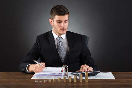 Young Businessman Calculating Tax With Stack Of Coins At Deskの写真素材