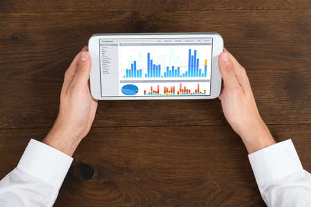 High Angle View Of Businessperson With Mobile Phone Showing Graph At Desk